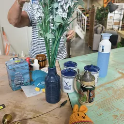 Tafelscène met diverse decoratieve voorwerpen: vaas met kunstbloemen, blikken, keramiek en een fles op een groene ondergrond