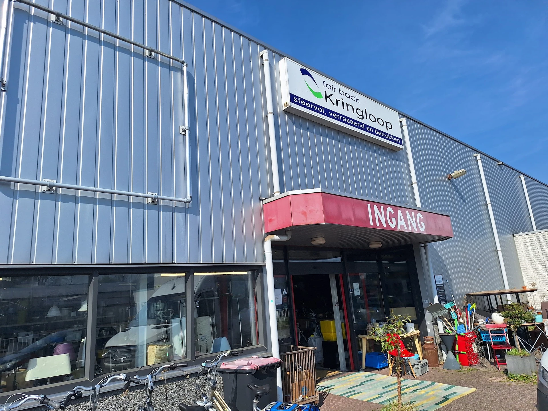 Gevel van kringloopwinkel met rode INGANG-luifel en grijze golfplaten wand