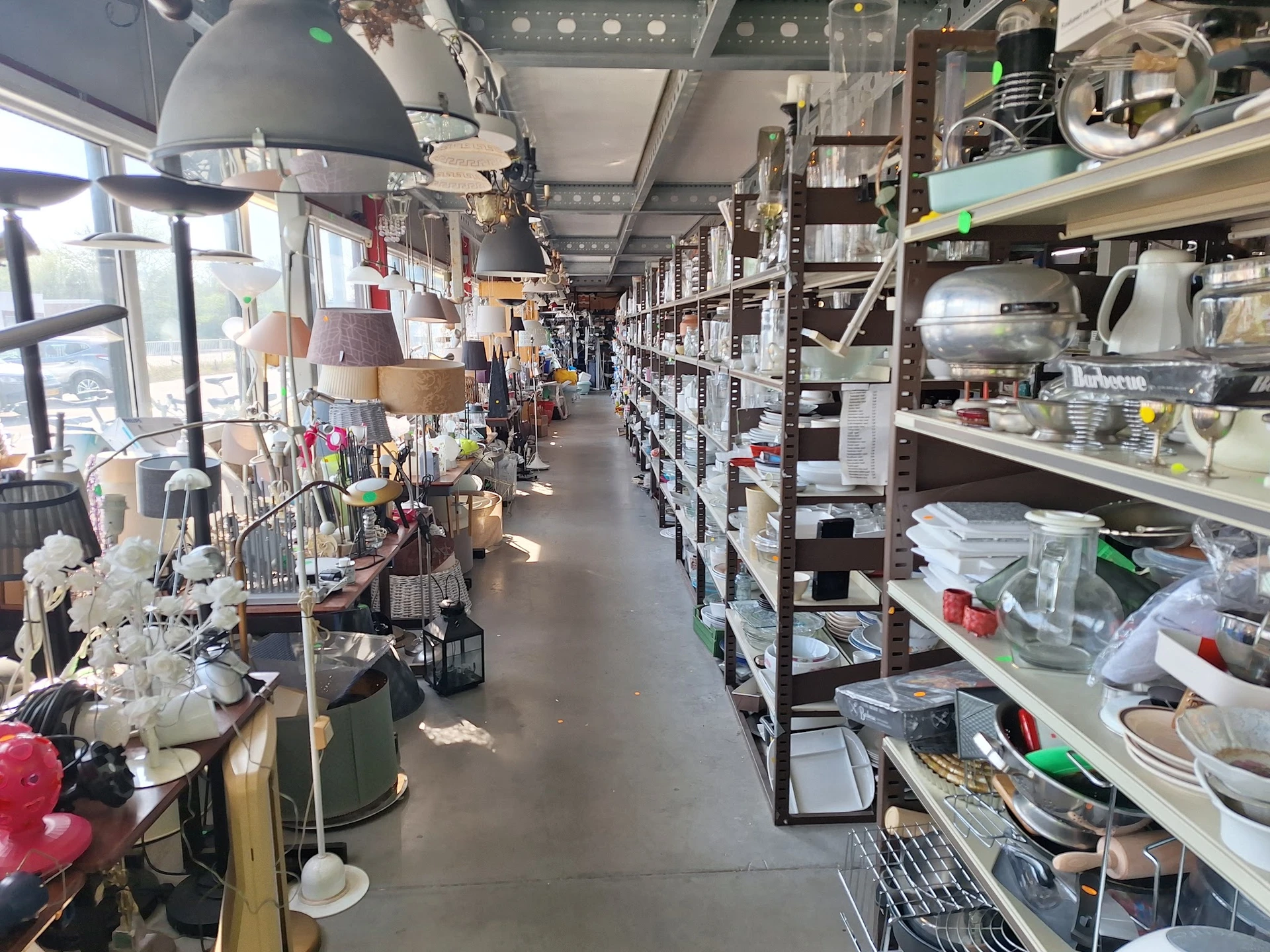 Lange gang in kringloopwinkel met stellingen vol spullen aan beide zijden en hangende artikelen