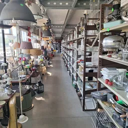 Lange gang in kringloopwinkel met stellingen vol spullen aan beide zijden en hangende artikelen