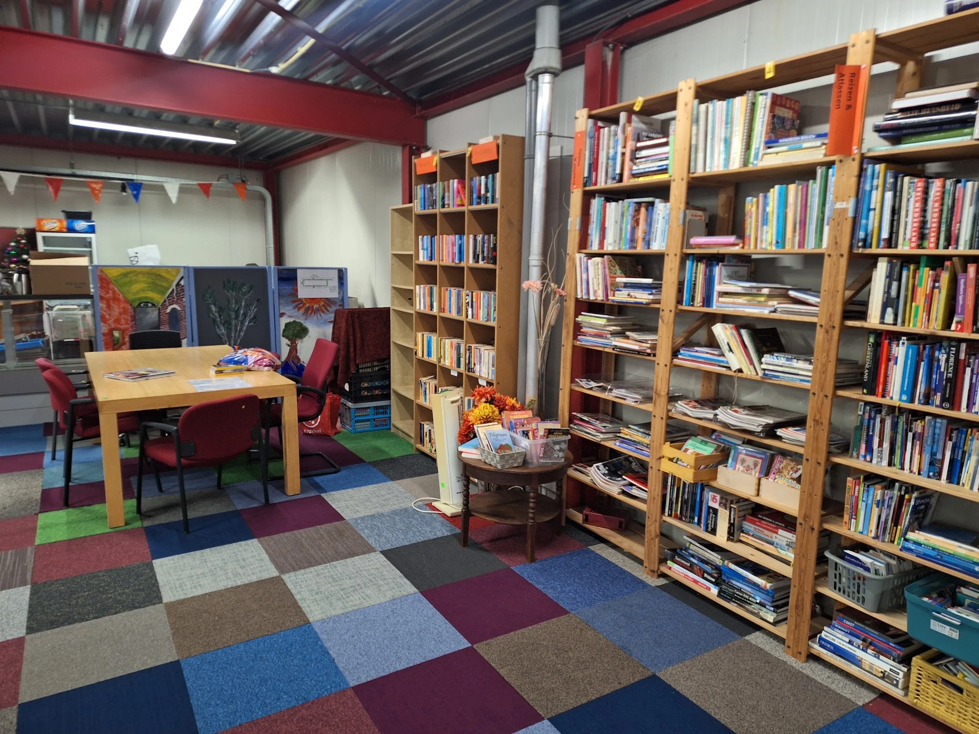 Binnenaanzicht van kringloopwinkel met houten boekenkasten vol boeken, houten tafel met stoelen, gekleurde vloer en rode mure