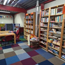 Binnenaanzicht van kringloopwinkel met houten boekenkasten vol boeken, houten tafel met stoelen, gekleurde vloer en rode mure