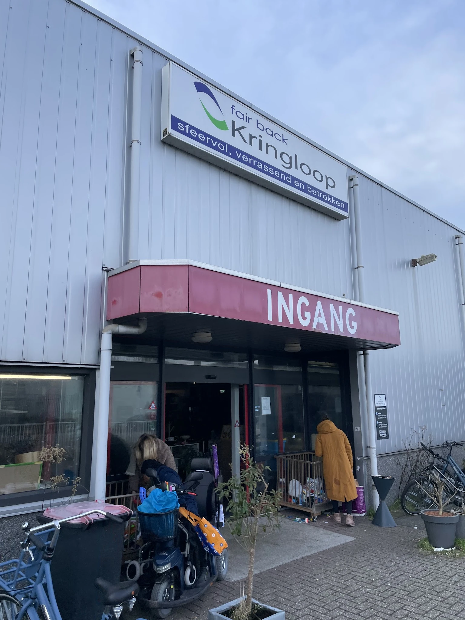 Ingang van kringloopwinkel met rode luifel en witte golfwanden, enkele bezoekers bij deur