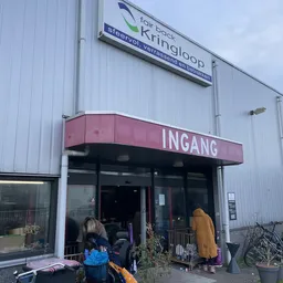 Ingang van kringloopwinkel met rode luifel en witte golfwanden, enkele bezoekers bij deur