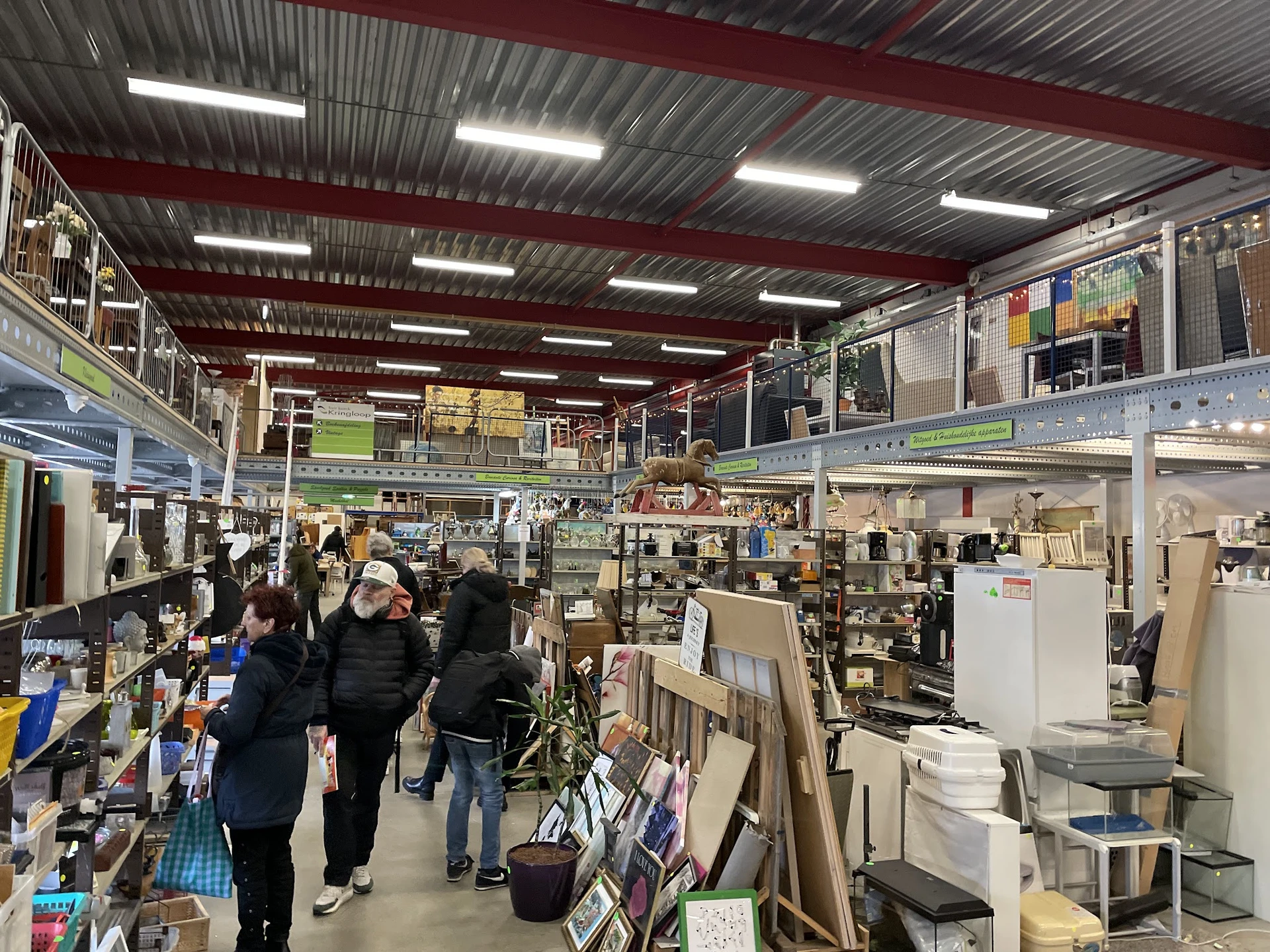 Overzicht van de winkelvloer met stellingen, rekken en uitgestalde spullen in een grote hal met rode balken