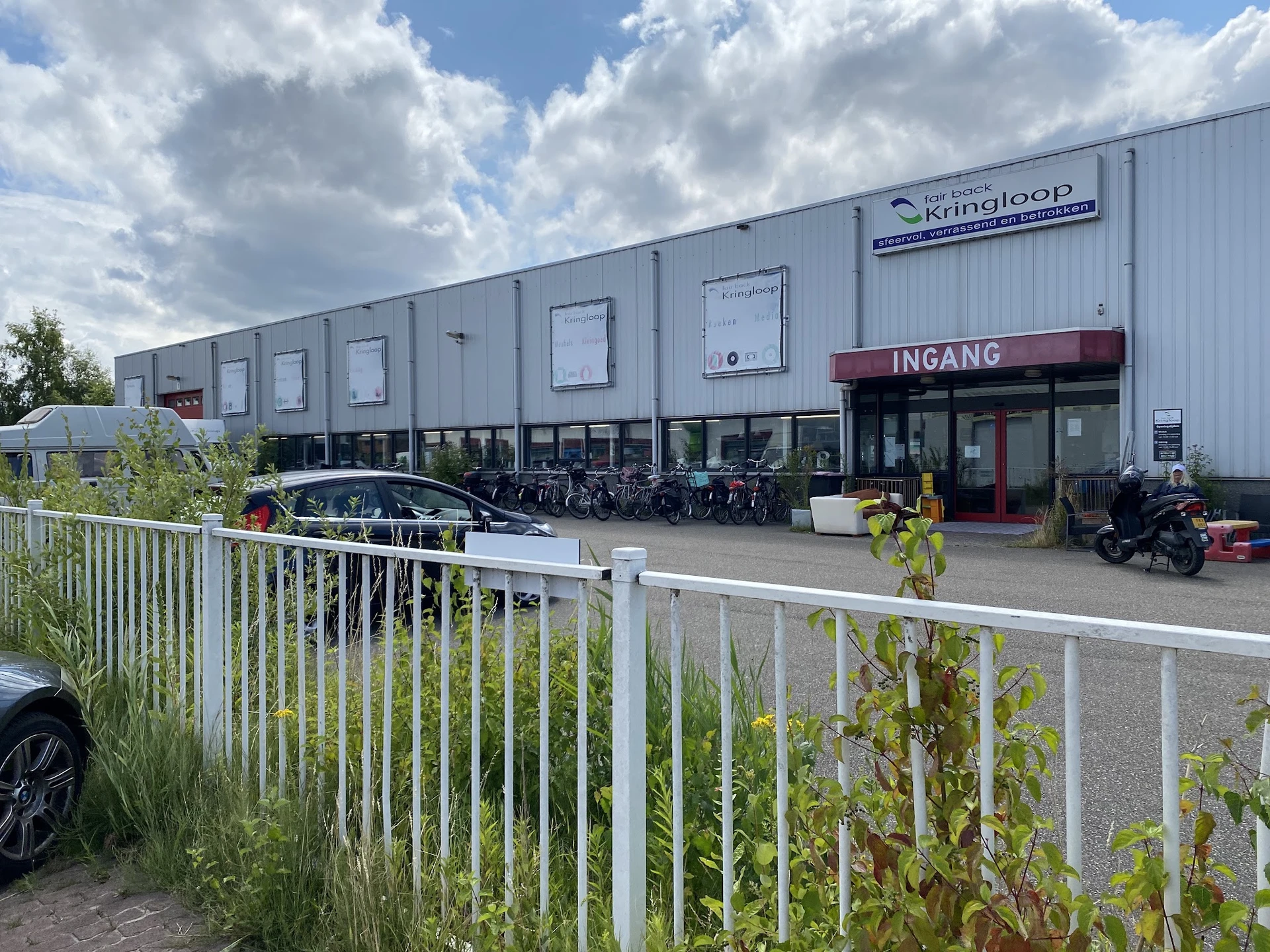 Grijze kunststof gevel van kringloopwinkel met INGANG-bord, geparkeerde auto's en motoren ervoor