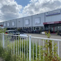 Grijze kunststof gevel van kringloopwinkel met INGANG-bord, geparkeerde auto's en motoren ervoor