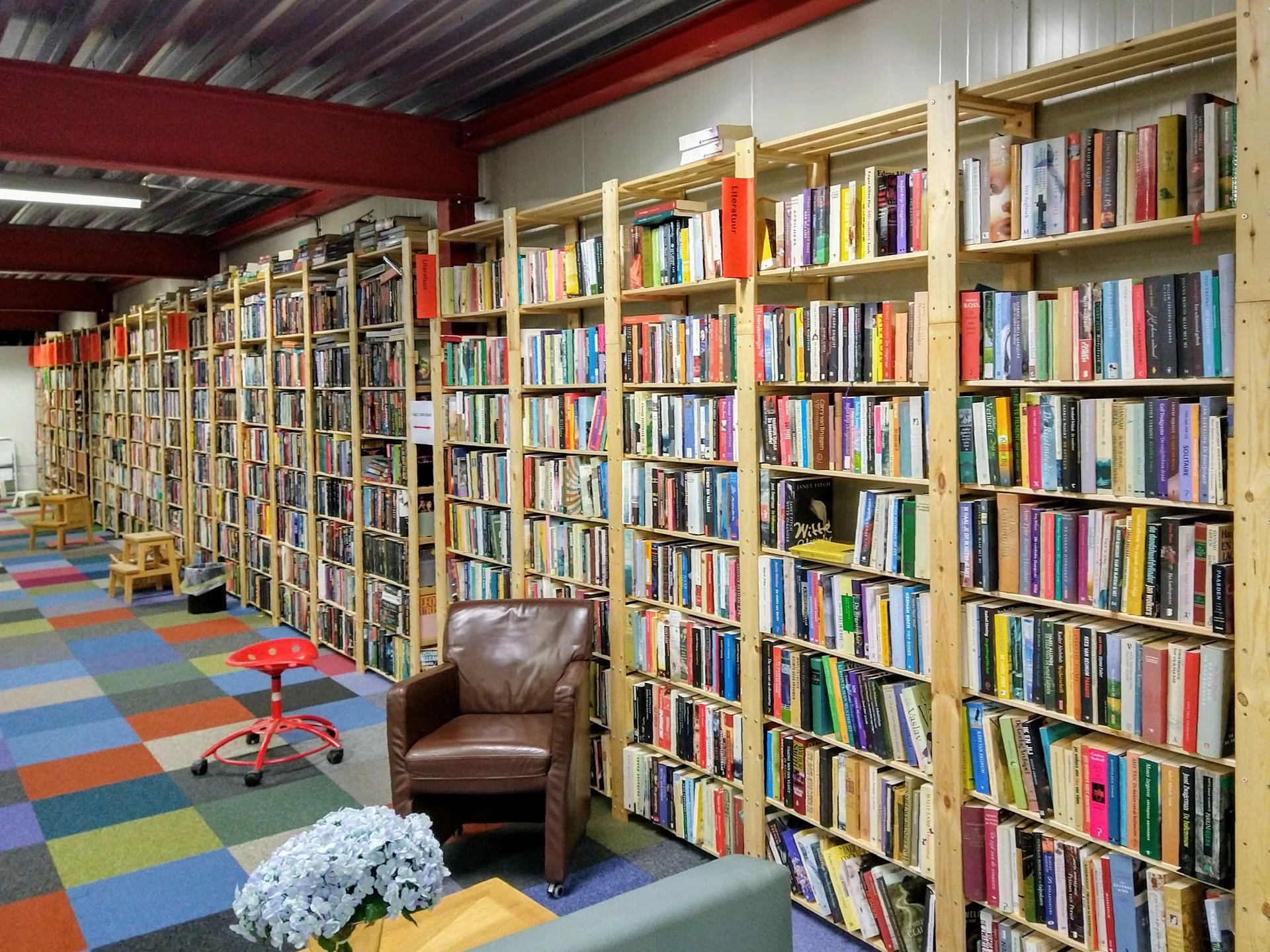 Binnenzicht van kringloopwinkel met uitgebreide boekenkasten langs de wanden, kleurrijke tegelvloer, bruine fauteuil en witte