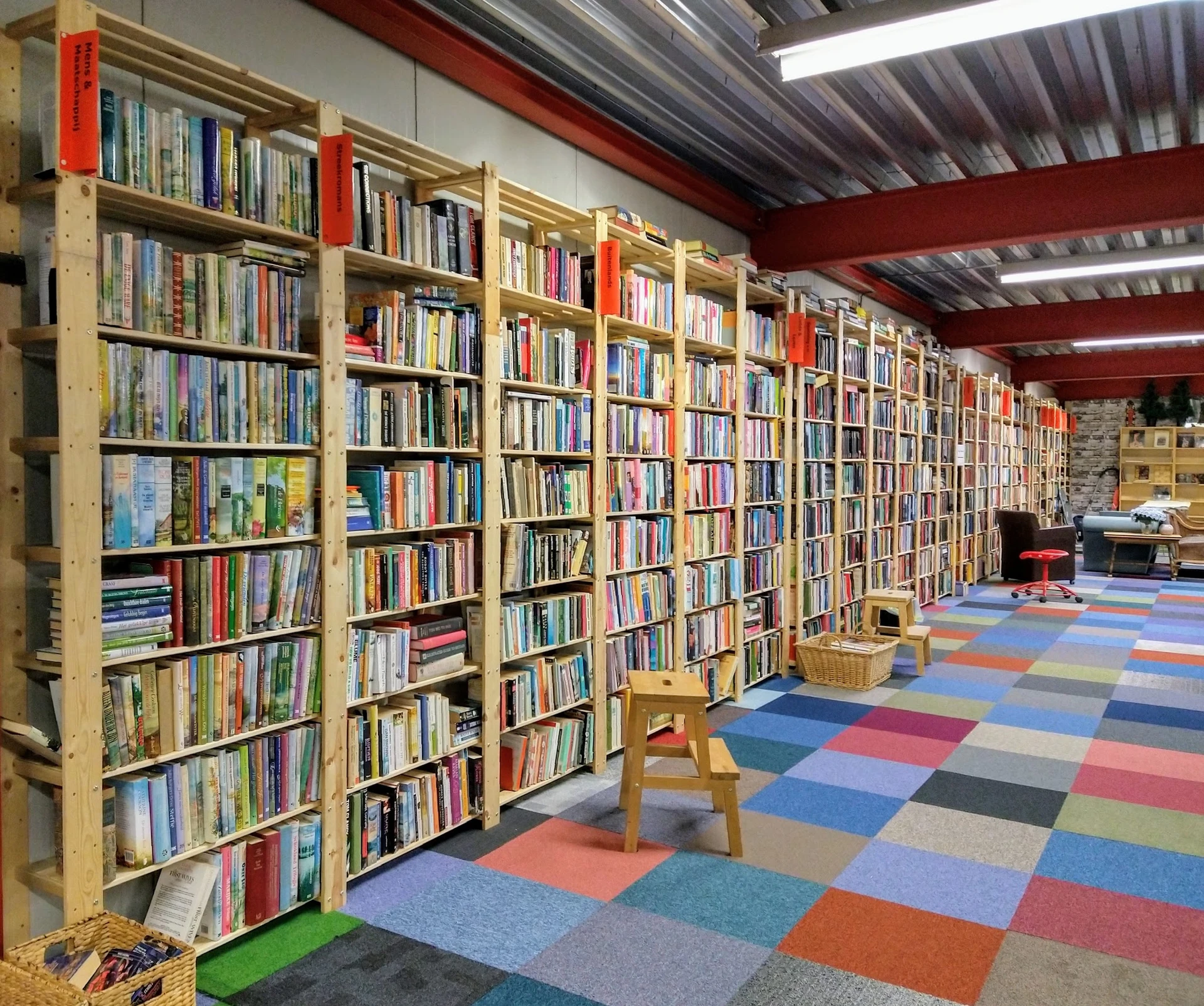 Binnenaanzicht van kringloopwinkel met houten boekenkasten vol tweedehands boeken, gekleurde tegelvloer en rode plafondspante