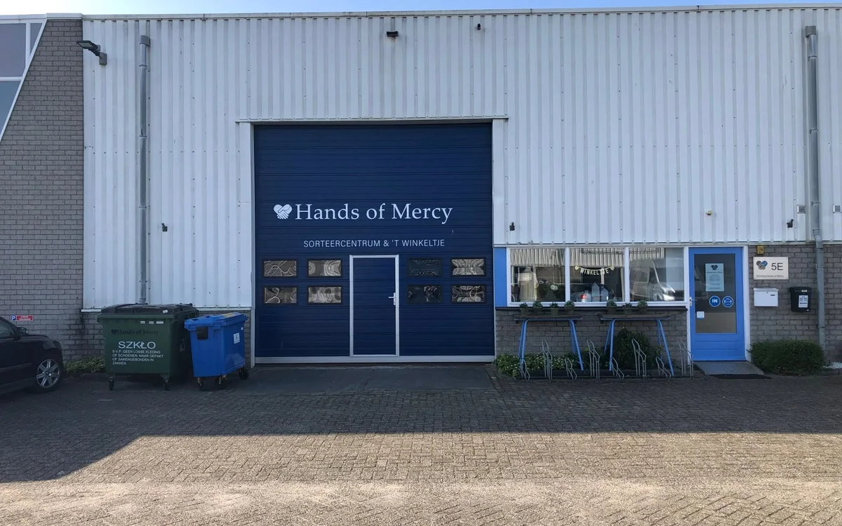 Witte bedrijfspand met donkerblauwe gevel en logo van kringloopwinkel, ingang met glazen deuren, parkeerplaats ervoor