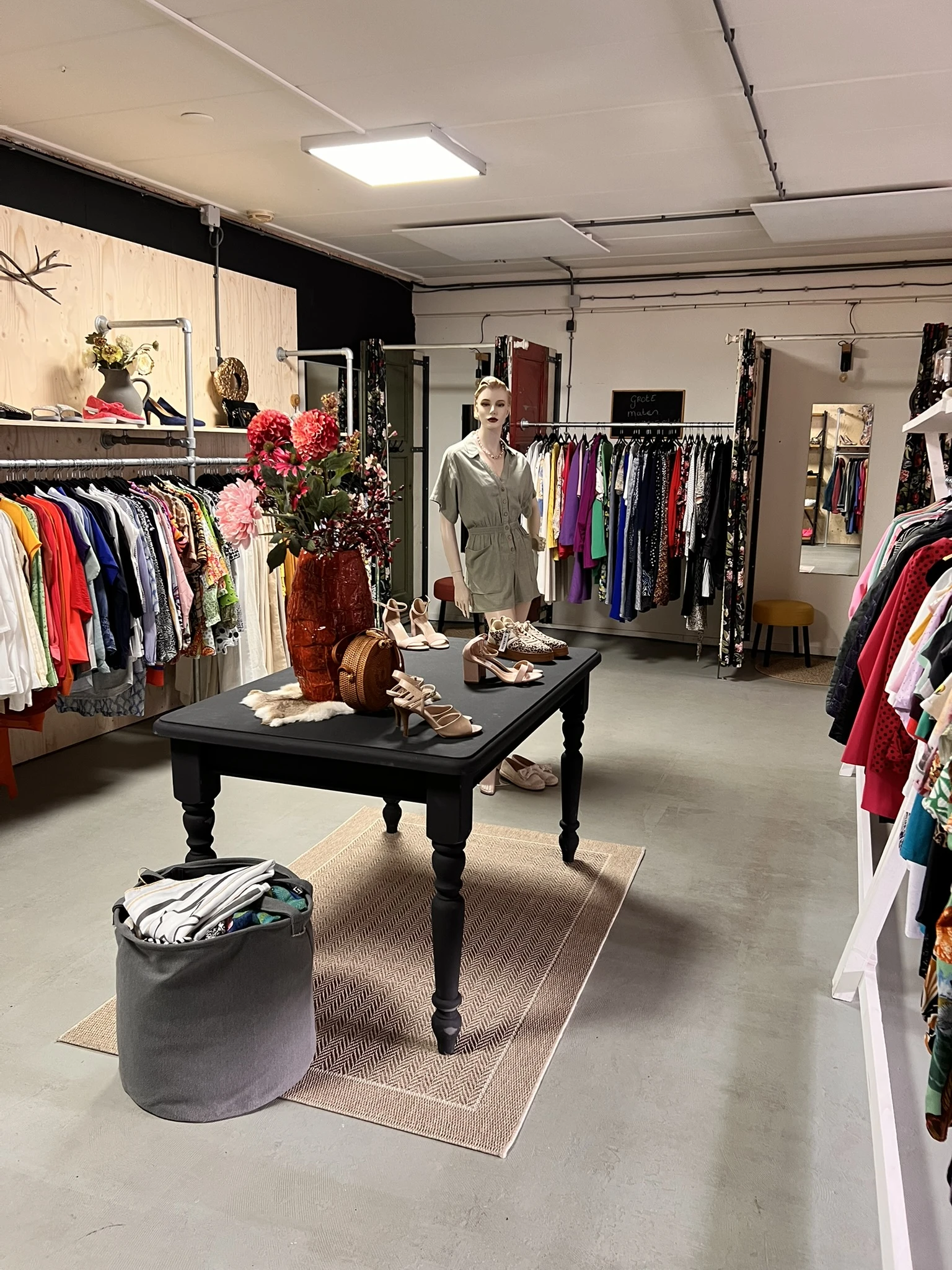 Winkelvloer met kledingrekken, zwarte tafel met schoenen en accessoires, bloemen, en vrouw in grijs shirt