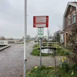Wegwijzers en richtingsborden bij een gebouw met een klein bruggetje over een sloot