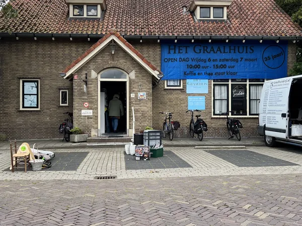 Gevel van kringloopwinkel Het Graalhuis met blauwe openingstijdenbord, ingang en fietsen ervoor
