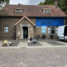 Gevel van kringloopwinkel Het Graalhuis met blauwe openingstijdenbord, ingang en fietsen ervoor