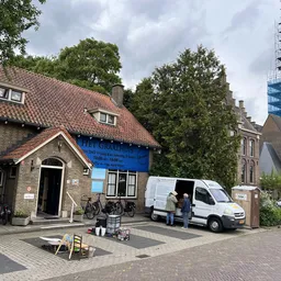 Voorgevel van een kringloopwinkel met rode dakpannen, blauwe deur en witte bestelbus geparkeerd voor de ingang