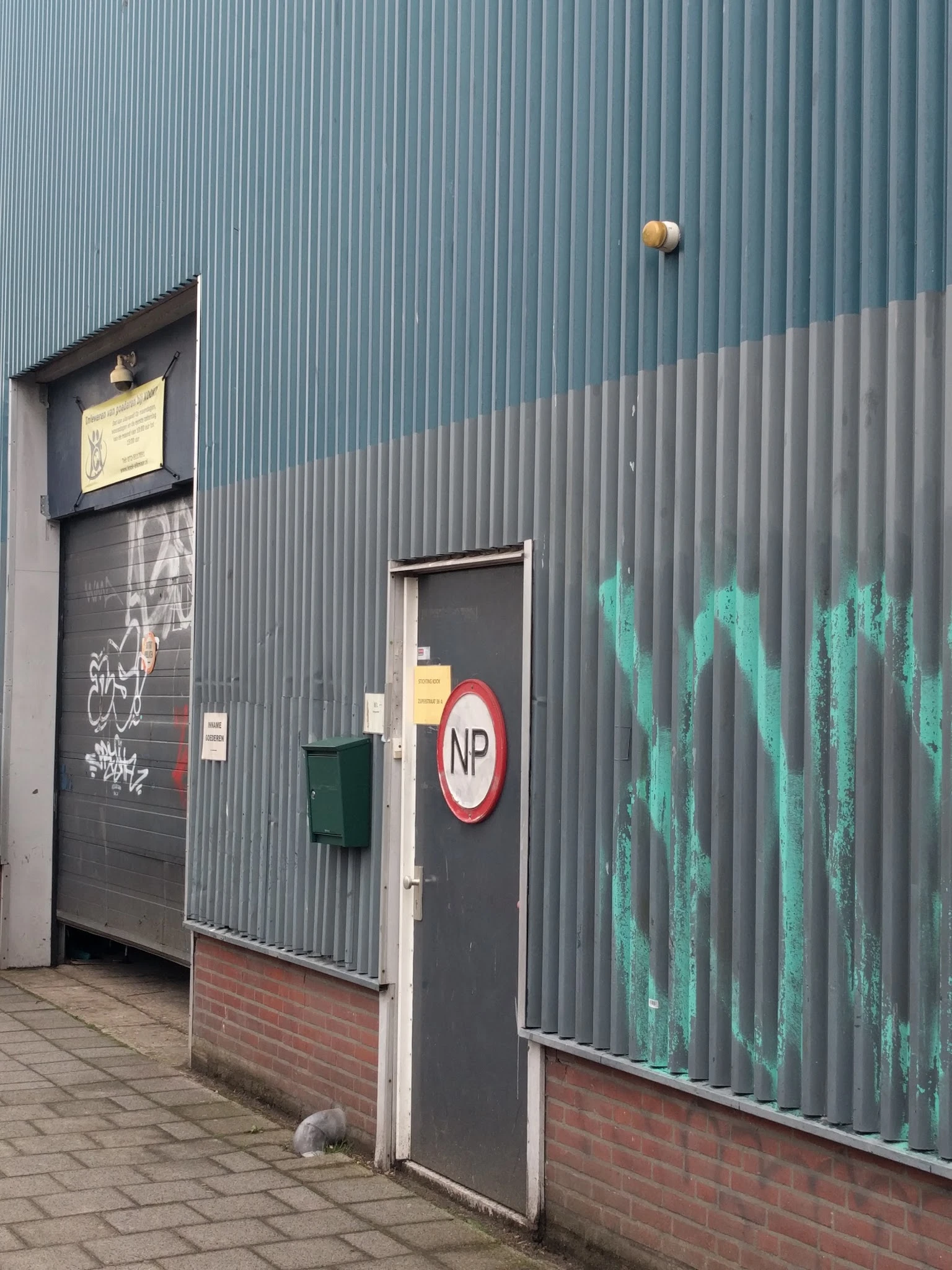 Zijkant van een grijzen golfplaten gebouw met een deur, NP-bord en groene graffiti