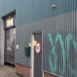 Zijkant van een grijzen golfplaten gebouw met een deur, NP-bord en groene graffiti