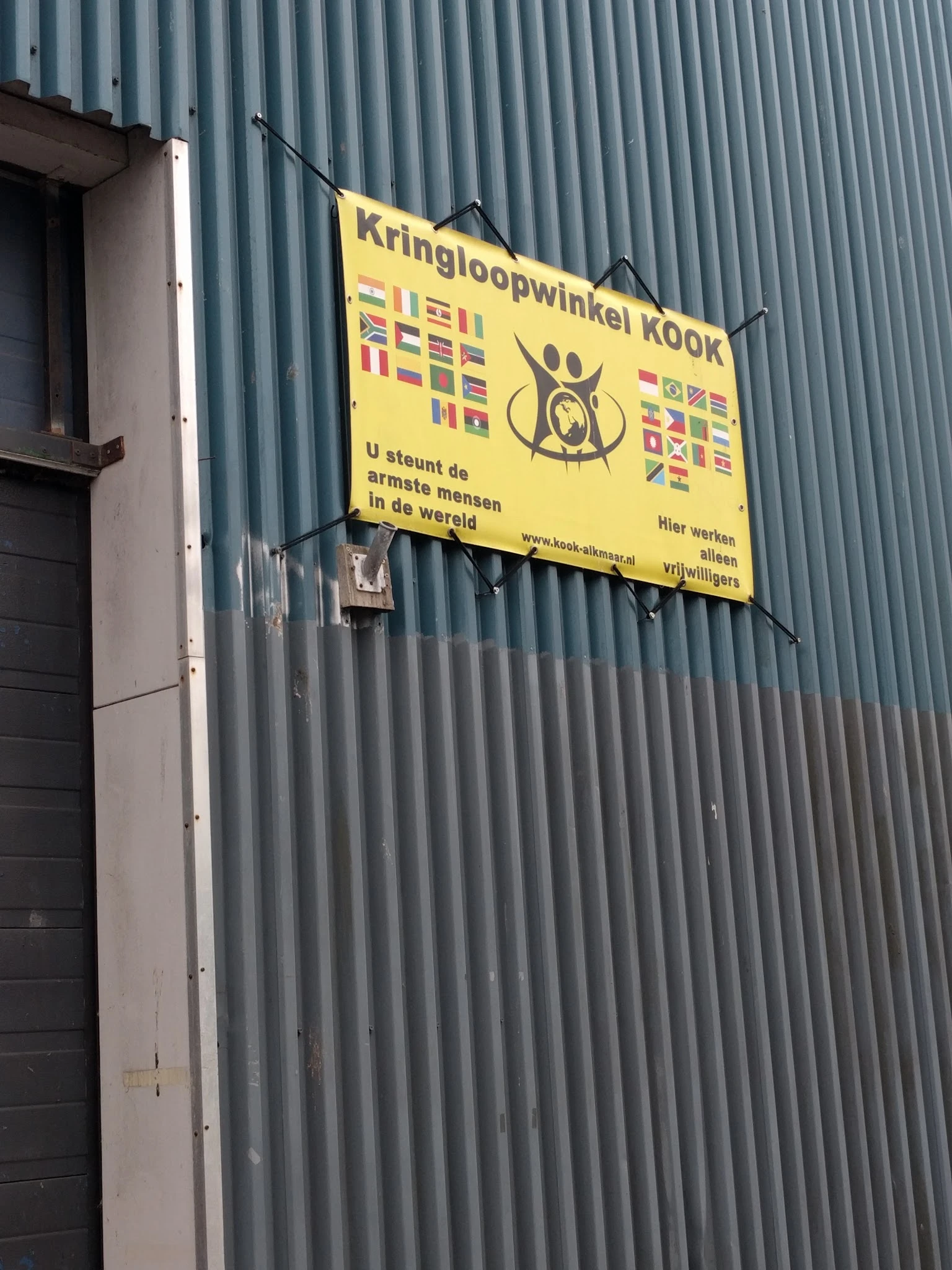 Grijze golfplaten gevel met geel uithangbord van Stichting Kook met internationale vlaggen