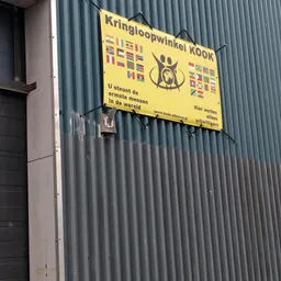 Grijze golfplaten gevel met geel uithangbord van Stichting Kook met internationale vlaggen