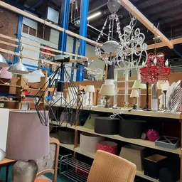 Diverse lampen en kroonluchters in verschillende stijlen op een tafel en aan het plafond in een kringloopwinkel