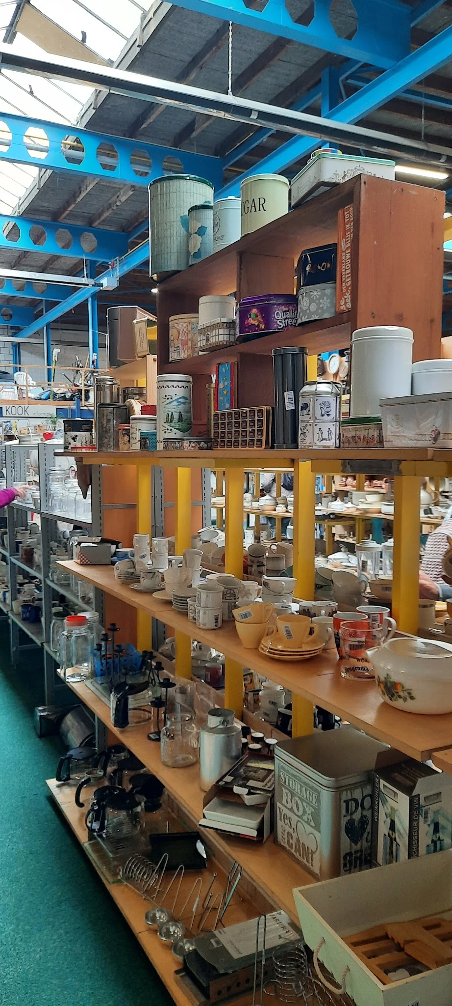 Servies, bekers en aardewerk uitgestald op toonbank in kringloopwinkel met gele stellingkasten op achtergrond