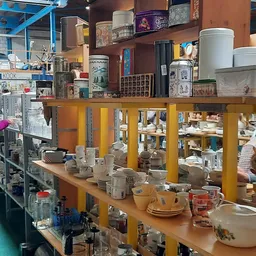 Servies, bekers en aardewerk uitgestald op toonbank in kringloopwinkel met gele stellingkasten op achtergrond