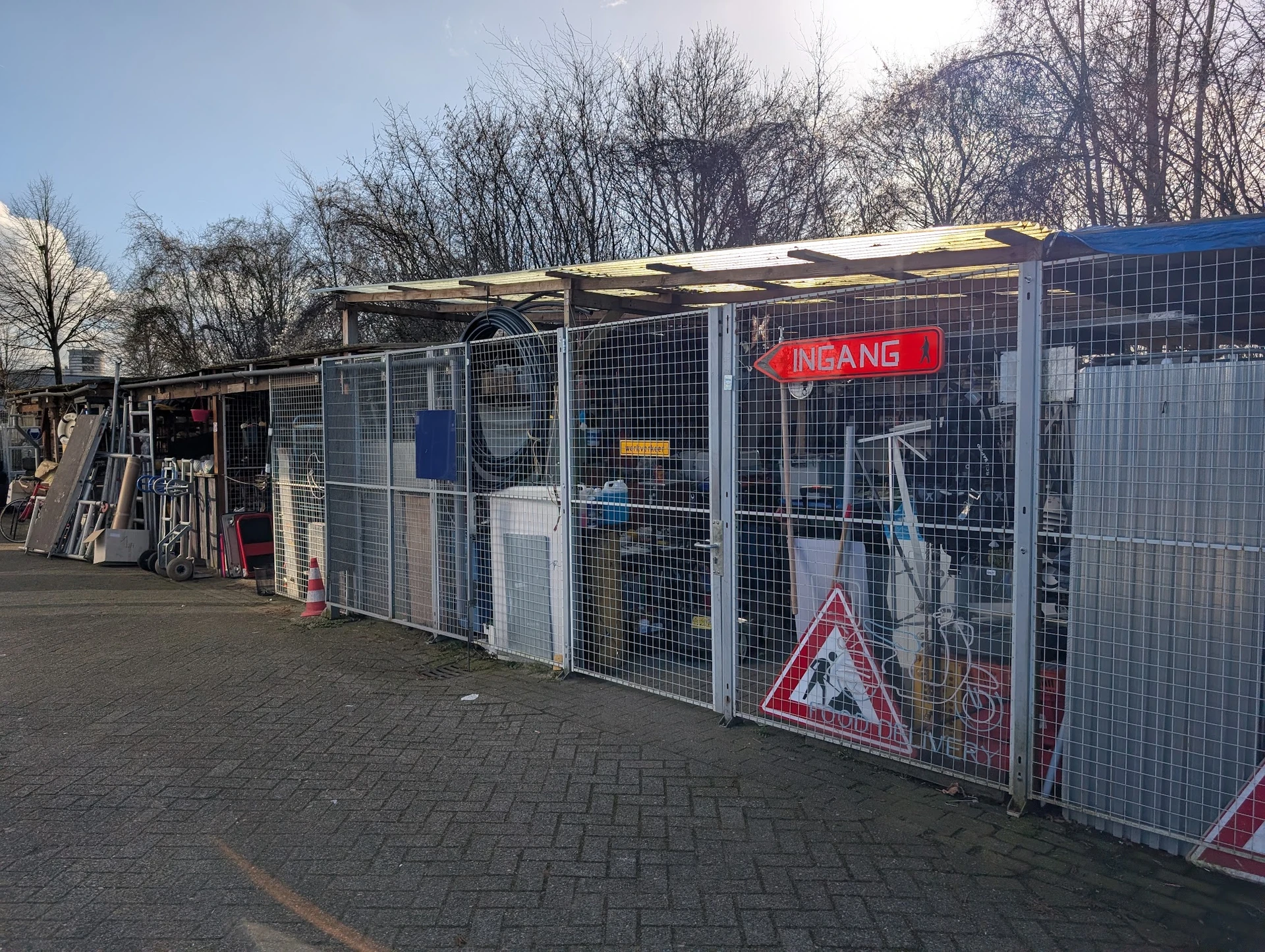 Voorkant van een kringloopwinkel met grijze metalen omheining, rode INGANG-verlichting en zonnepanelen op het dak
