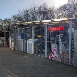 Voorkant van een kringloopwinkel met grijze metalen omheining, rode INGANG-verlichting en zonnepanelen op het dak