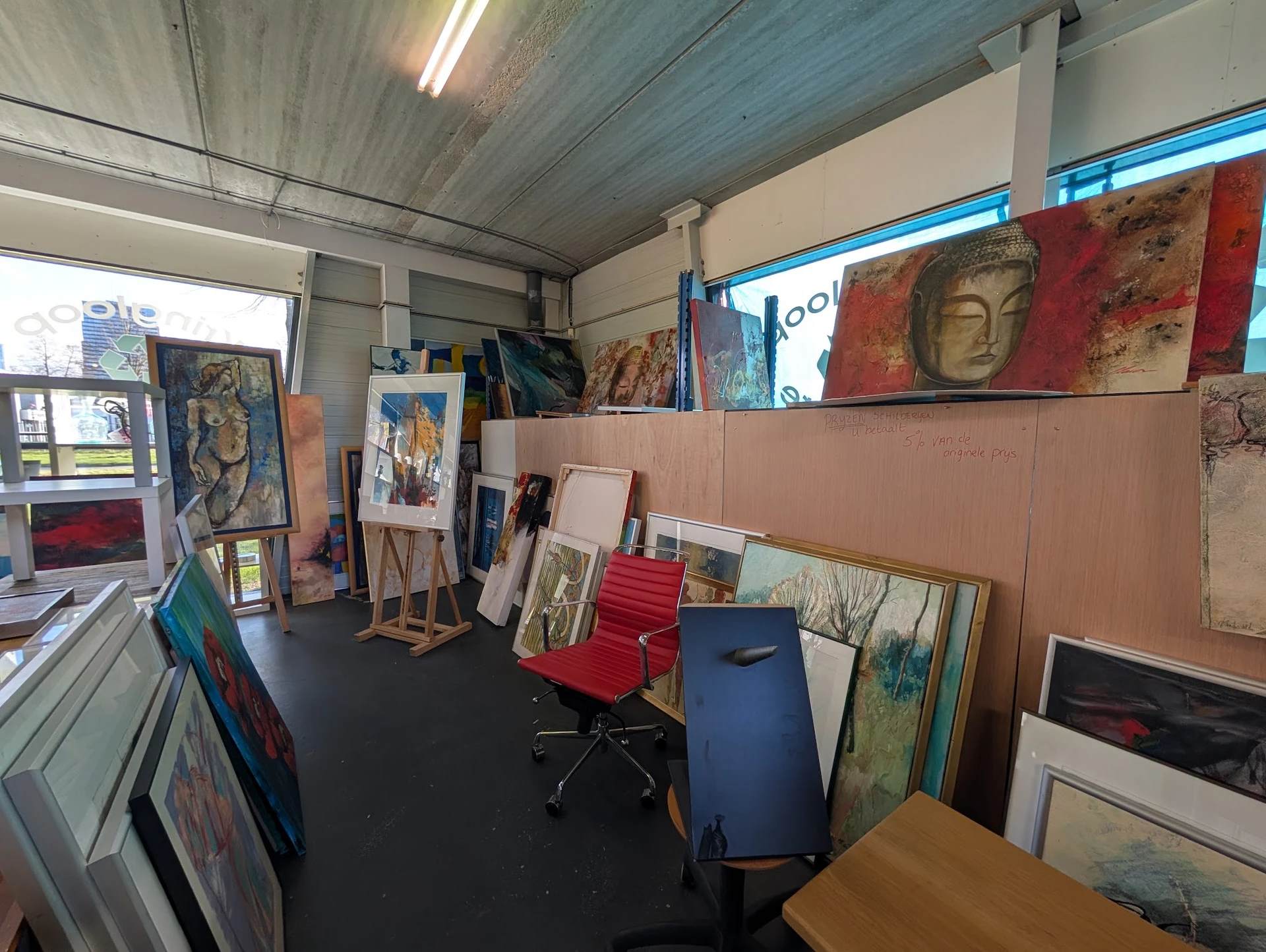 Werkruimte in kringloopwinkel met kunstwerken, schilderijen en frames tegen muren, stellingen met spullen en zwarte vloer