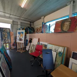 Werkruimte in kringloopwinkel met kunstwerken, schilderijen en frames tegen muren, stellingen met spullen en zwarte vloer
