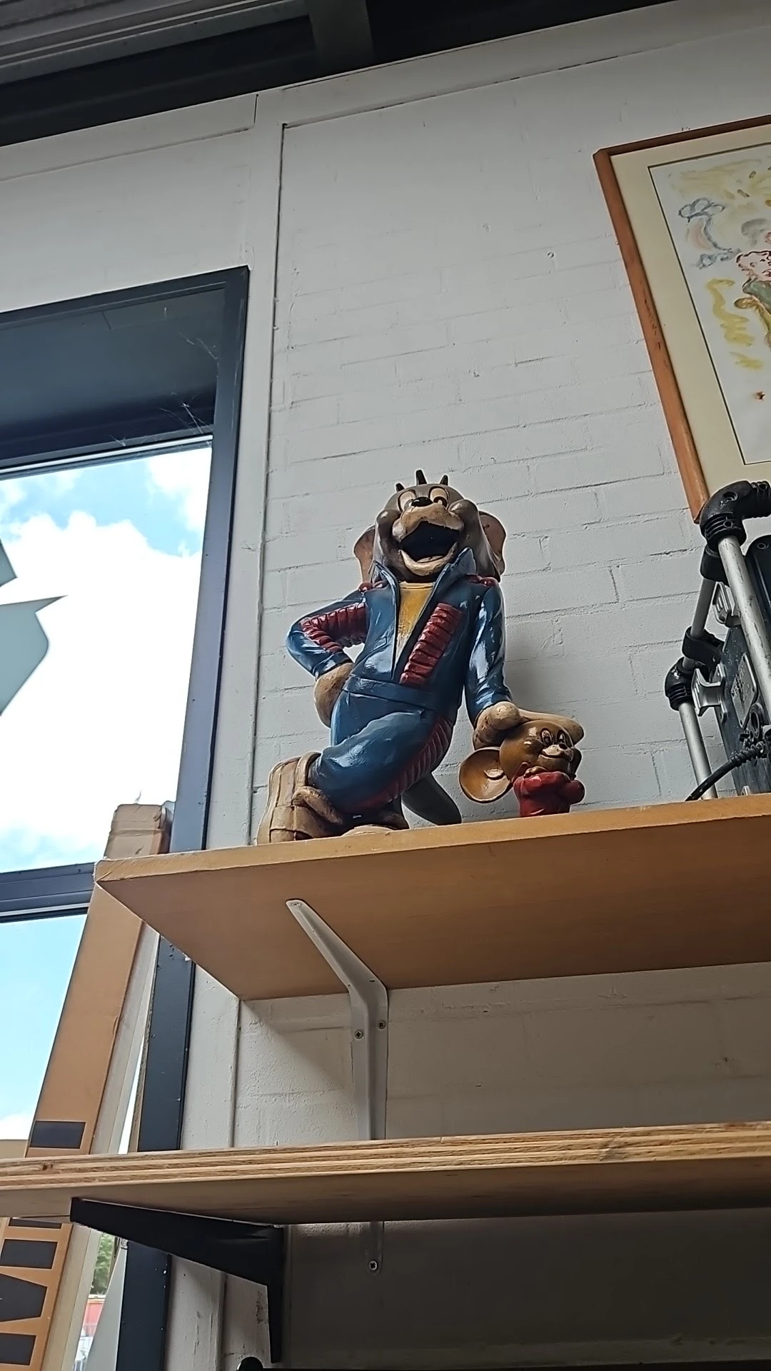 Groot houten beeldhouwwerk van een lachend dier in kleding op een houten balk buiten de winkel
