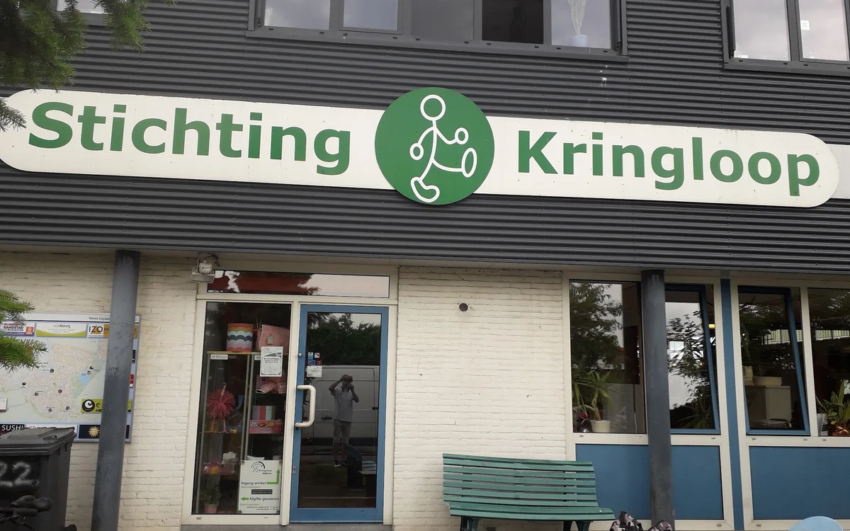 Voorgevel van Stichting Kringloop met groene letters, witte bakstenen onderkant en blauwe raamframes, groene zitbank op het t