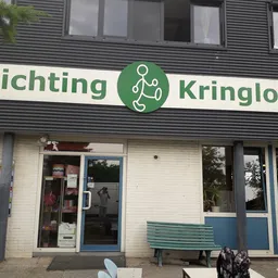 Voorgevel van Stichting Kringloop met groene letters, witte bakstenen onderkant en blauwe raamframes, groene zitbank op het t