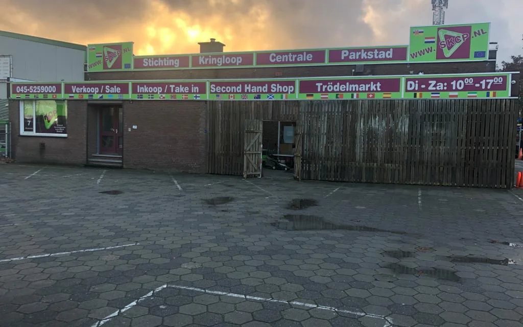Gevel van kringloopwinkel met groene uithangborden en donkere puien aan pleinen