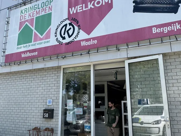Voorgevel van kringloopwinkel met witte kozijnen, reclameborden en persoon in deuropening