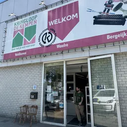 Voorgevel van kringloopwinkel met witte kozijnen, reclameborden en persoon in deuropening