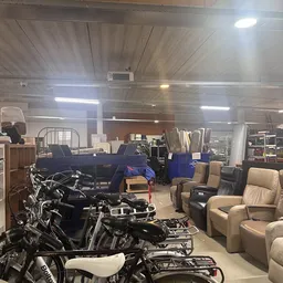 Groot magazijn met tweedehandsartikelen: motoren, een blauwe auto, meubels en diverse spullen