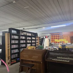 Binnenaanzicht van kringloopwinkel met cd/dvd-kasten, piano, muziekuitrusting en diverse spullen