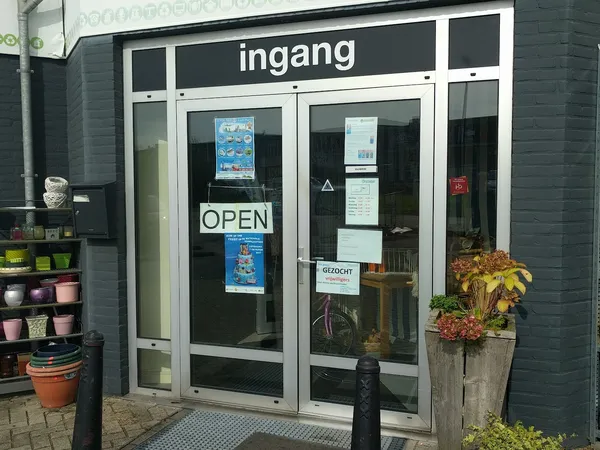 Glazen ingang van kringloopwinkel met 'ingang' opschrift, OPEN-bord en informatiepapieren, plantenbakken ernaast