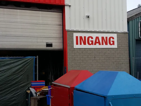 Voorgevel van kringloopwinkel met rood INGANG-bord, garagedeur en gekleurde containers