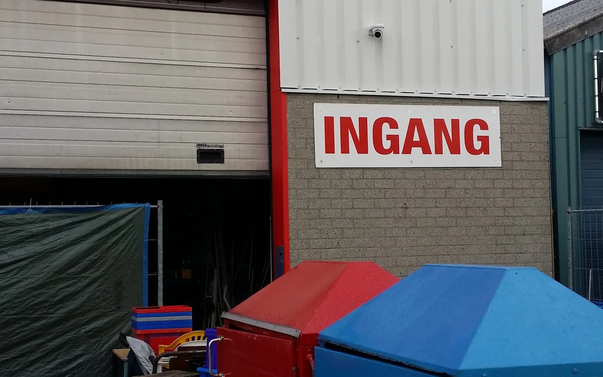 Voorgevel van kringloopwinkel met rood INGANG-bord, garagedeur en gekleurde containers
