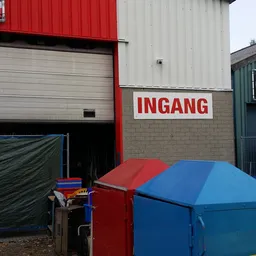 Voorgevel van kringloopwinkel met rood INGANG-bord, garagedeur en gekleurde containers