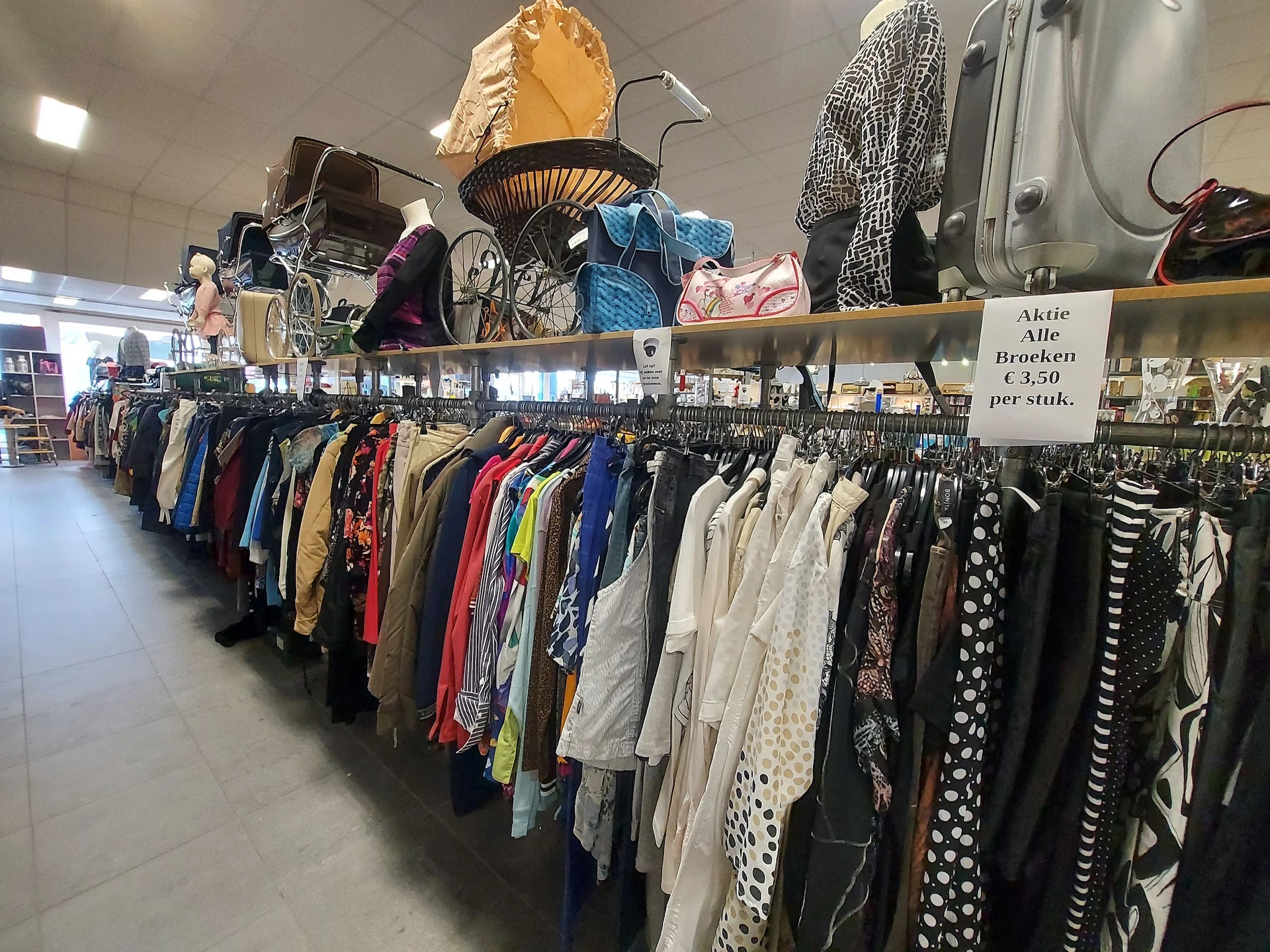 Lange rekken met tweedehands kleding in diverse kleuren en stijlen, met op de achtergrond displays met tassen en accessoires.