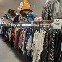 Lange rekken met tweedehands kleding in diverse kleuren en stijlen, met op de achtergrond displays met tassen en accessoires.