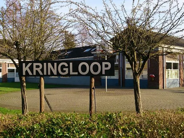 Gevel van kringloopwinkel met groot bord 'KRINGLOOP', bakstenen pui, ramen en twee jonge bomen ervoor