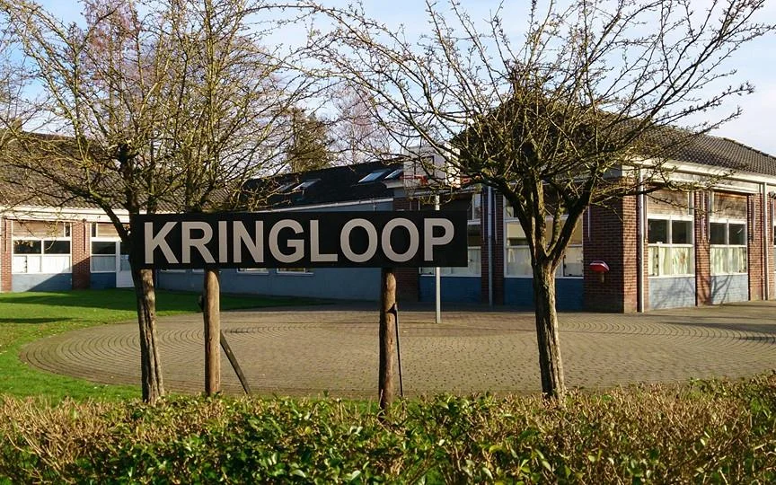 Gevel van kringloopwinkel met groot bord 'KRINGLOOP', bakstenen pui, ramen en twee jonge bomen ervoor