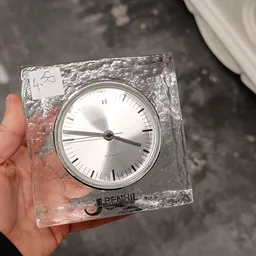 Close-up van een vierkante kwartswekker met witte wijzerplaat en zwarte wijzers, in plastic verpakking