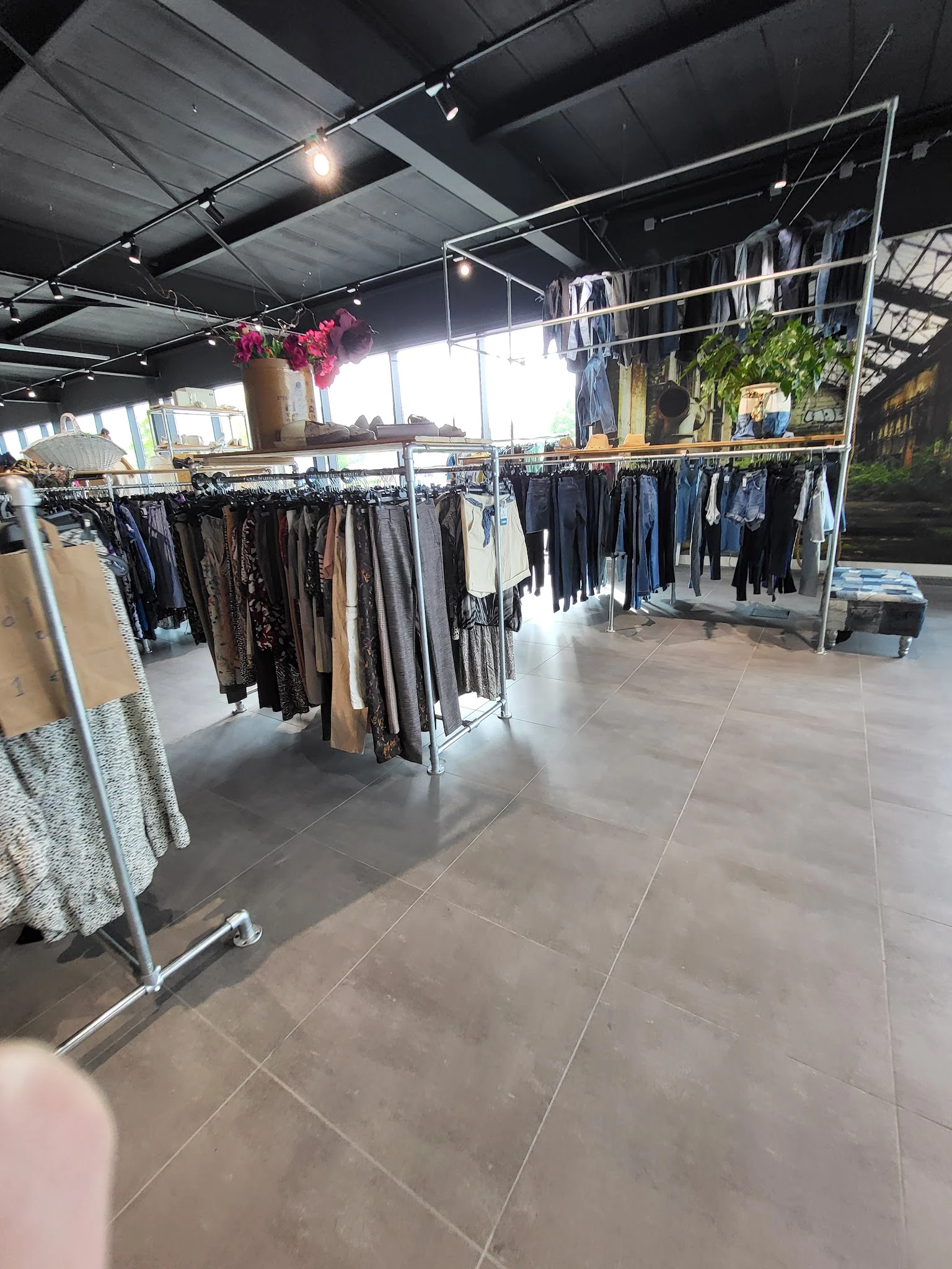 Winkelinterieur met kledingrekken vol tweedehands kleding, hangende bloemen en grote ramen met daglicht