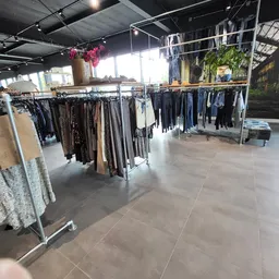 Winkelinterieur met kledingrekken vol tweedehands kleding, hangende bloemen en grote ramen met daglicht