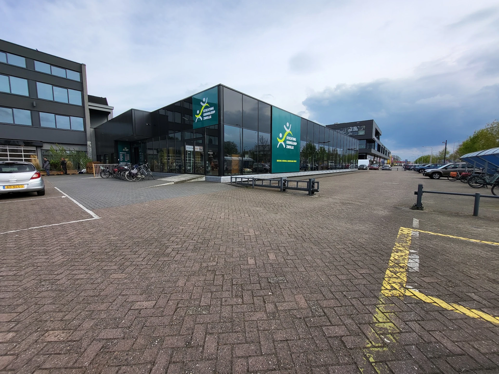 Voorgevel van kringloopwinkel met groene logo's en grote glazen pui aan een plein met parkeerplaatsen